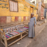 Älterer Mann in traditioneller Kleidung mit Besen und Sofa an der Strasse im Dorf Al Aqaltah bei Luxor, Ägypten, Afrika | Traditional dressed elderly man with broom and his Sofa on the street in Al Aqaltah village near Luxor, Egypt, Africa